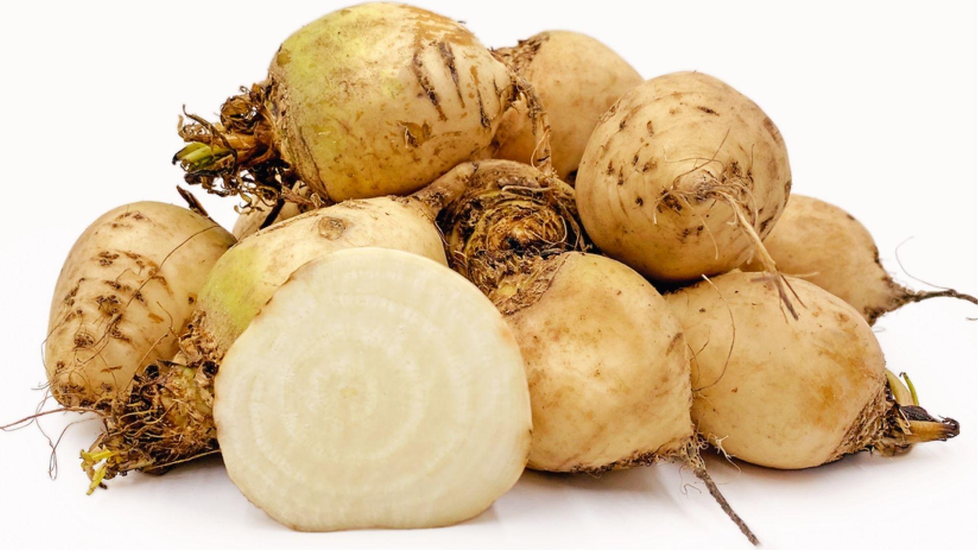 Pile of white beets with pale skin and one sliced open revealing creamy flesh on white background.