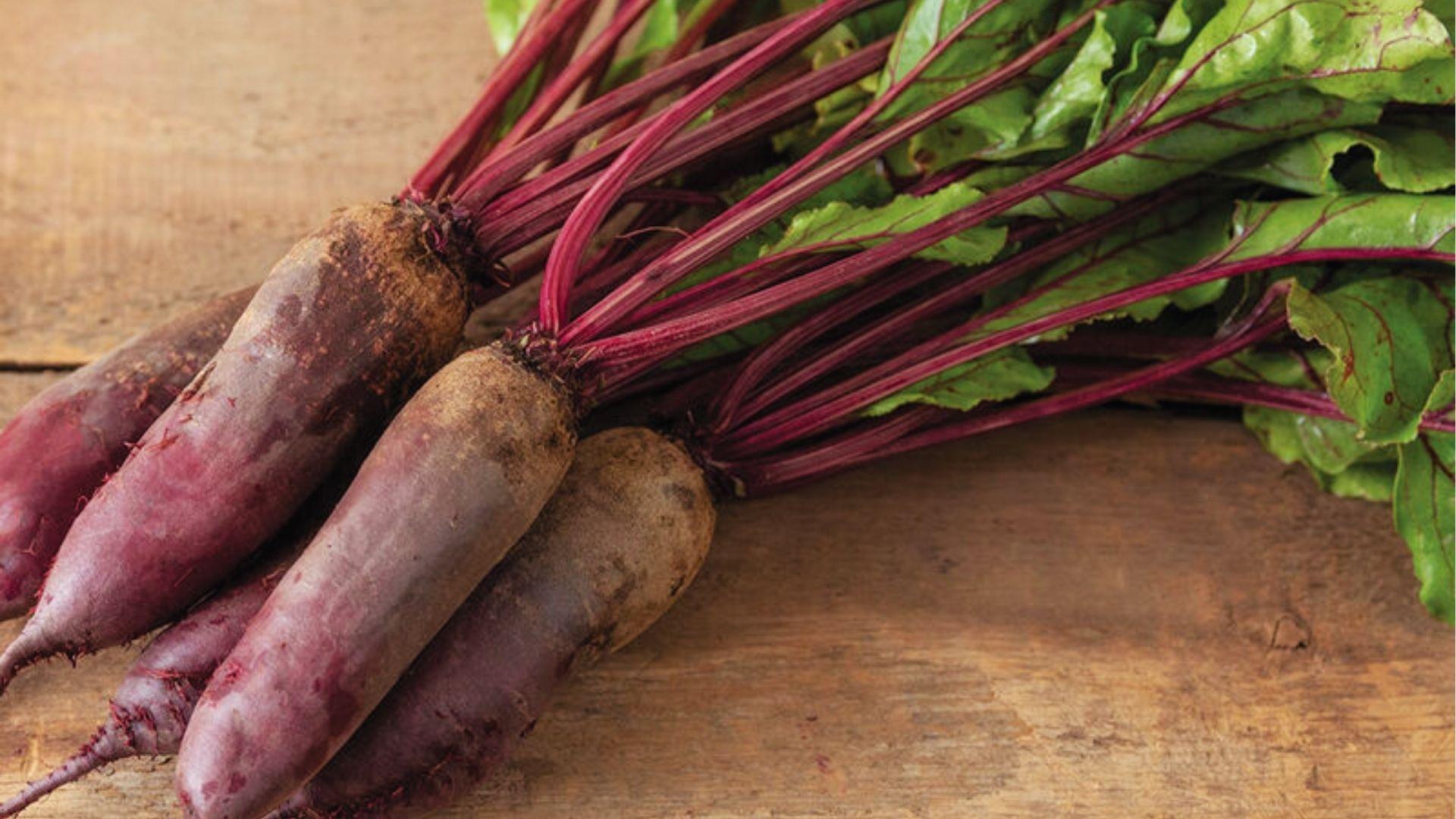 Long red beets with magenta stems and green leaves resting on a rustic wooden table.