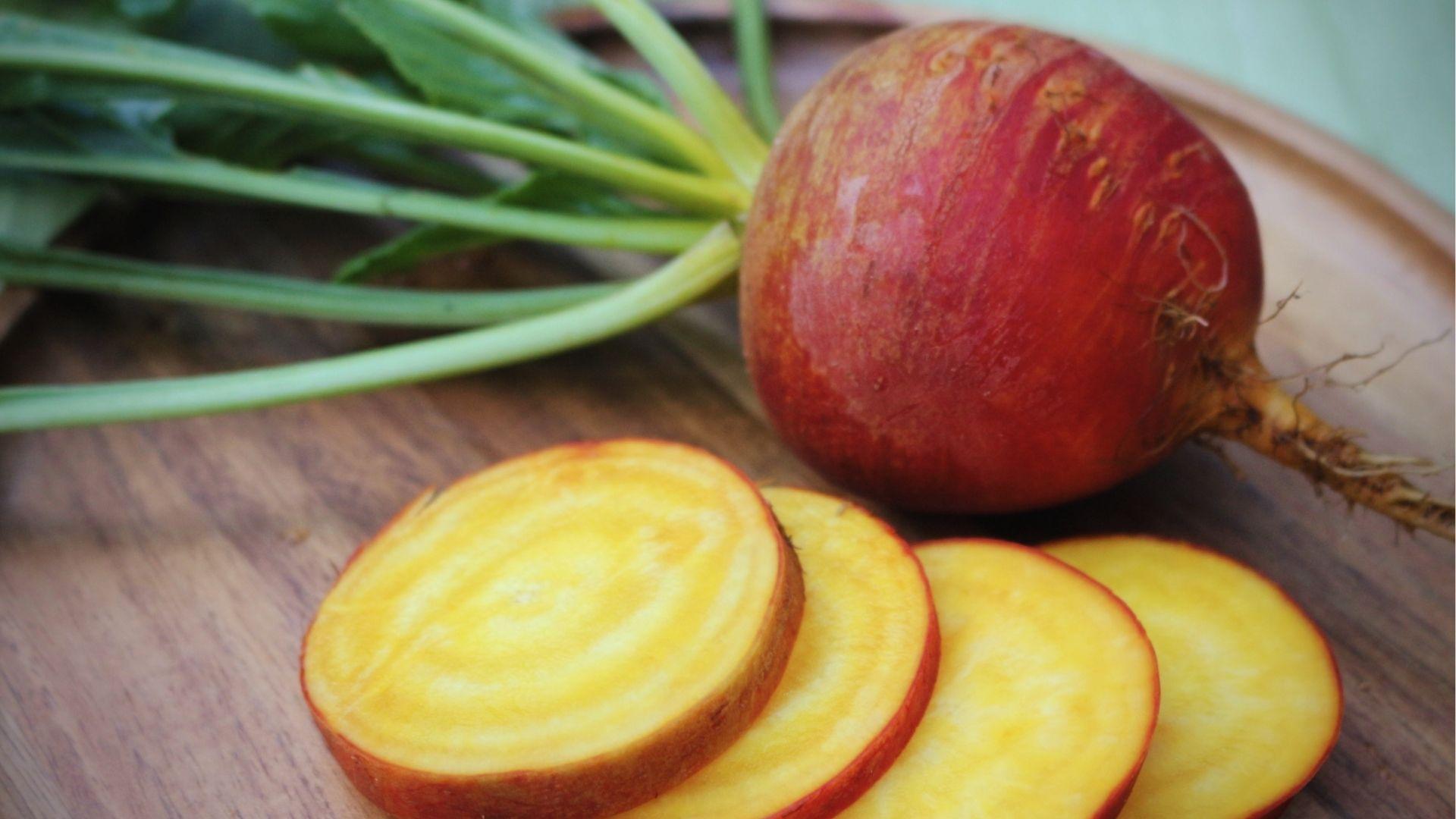 Large golden beet with red-orange skin and vivid yellow slices, green leaves, on a wood plate.