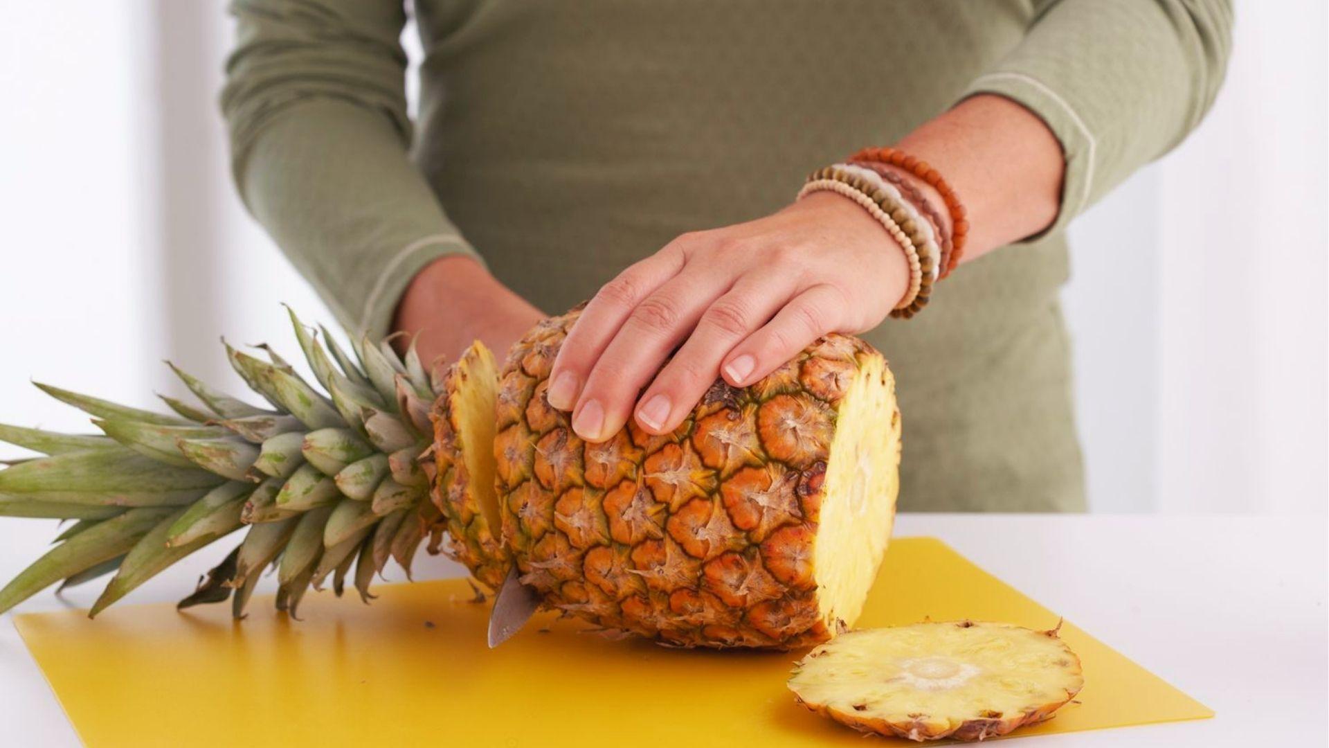 Person slicing top off ripe pineapple on yellow board, starting the propagation process