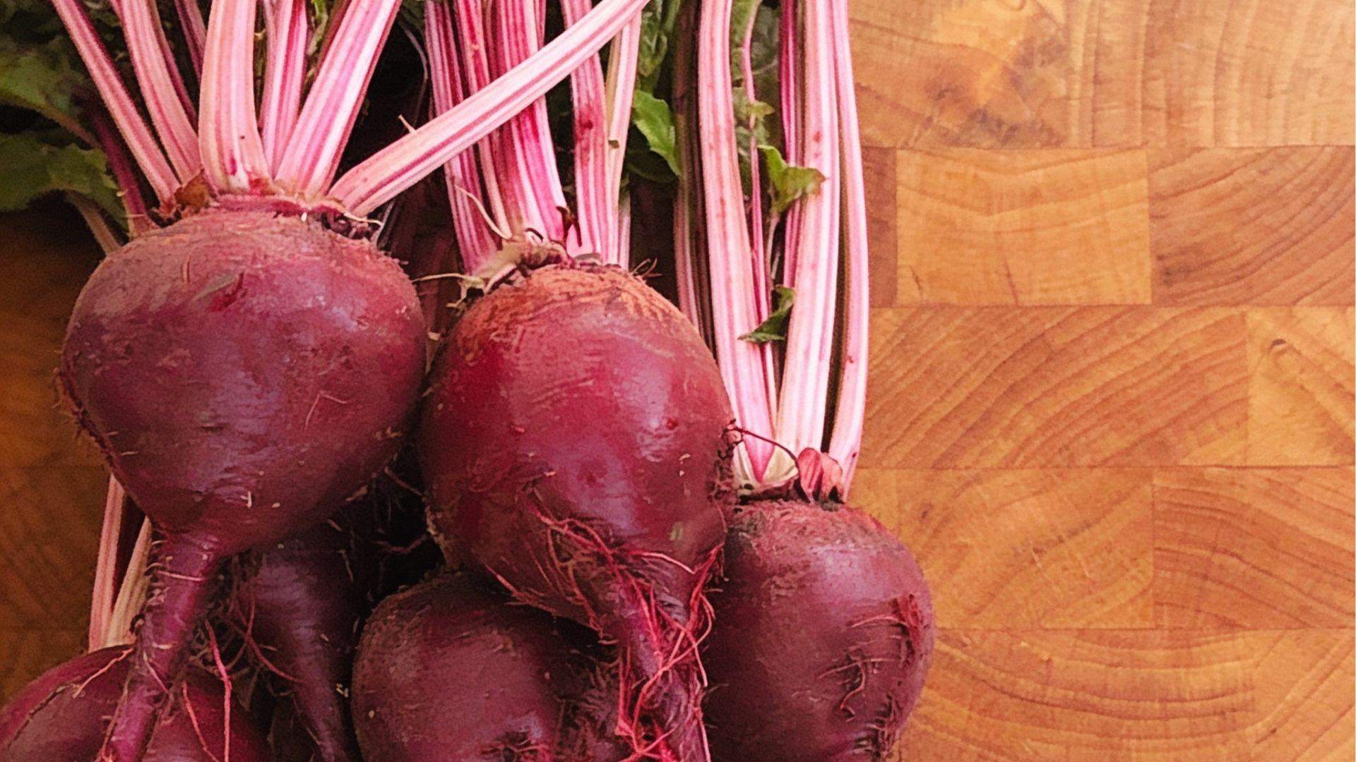 Bunch of round red beets with pink-striped stems and earthy skin on wooden cutting board closeup.