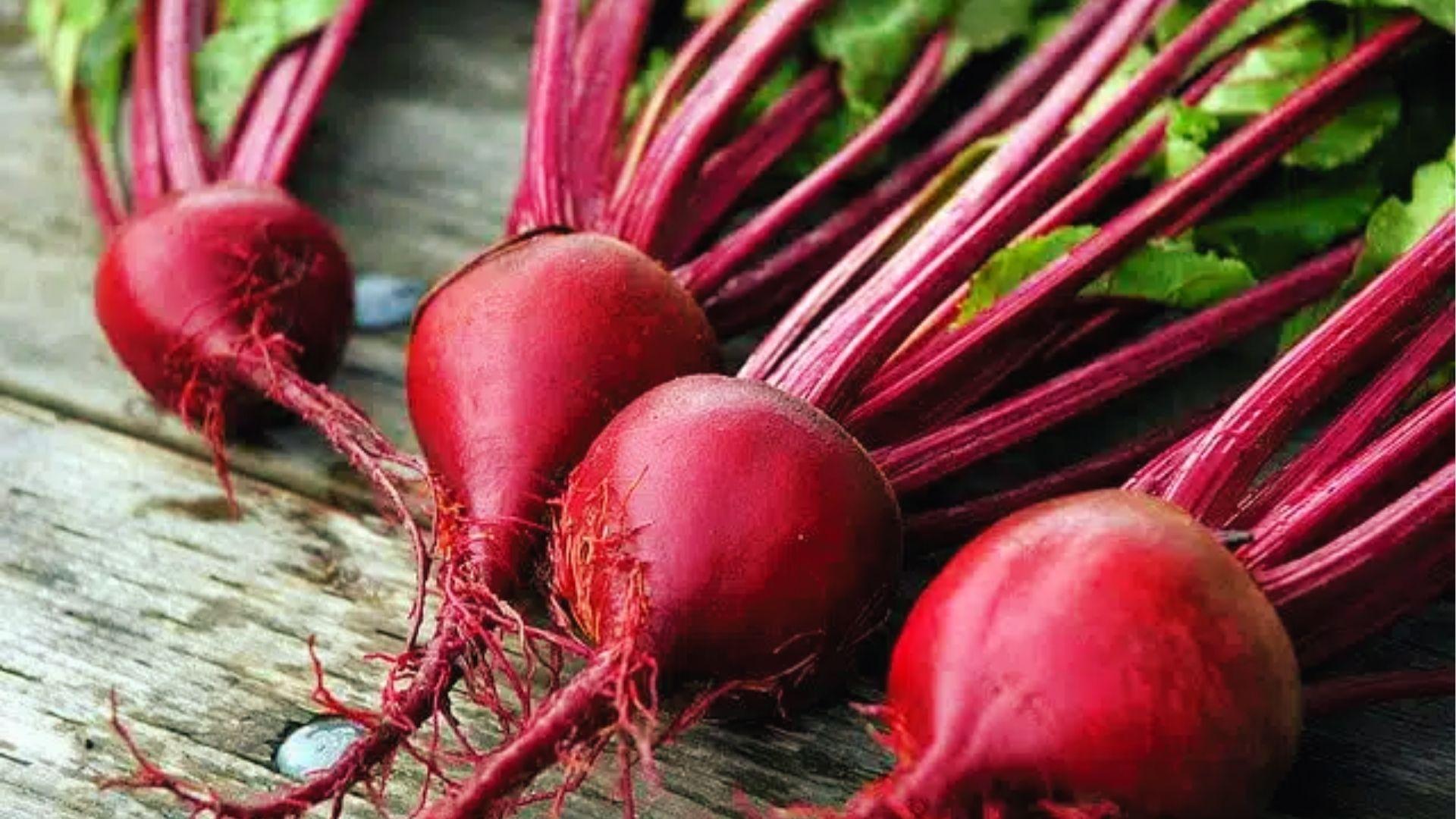 Four red beets with long crimson stems and thin roots arranged on weathered wood, a common beet type.