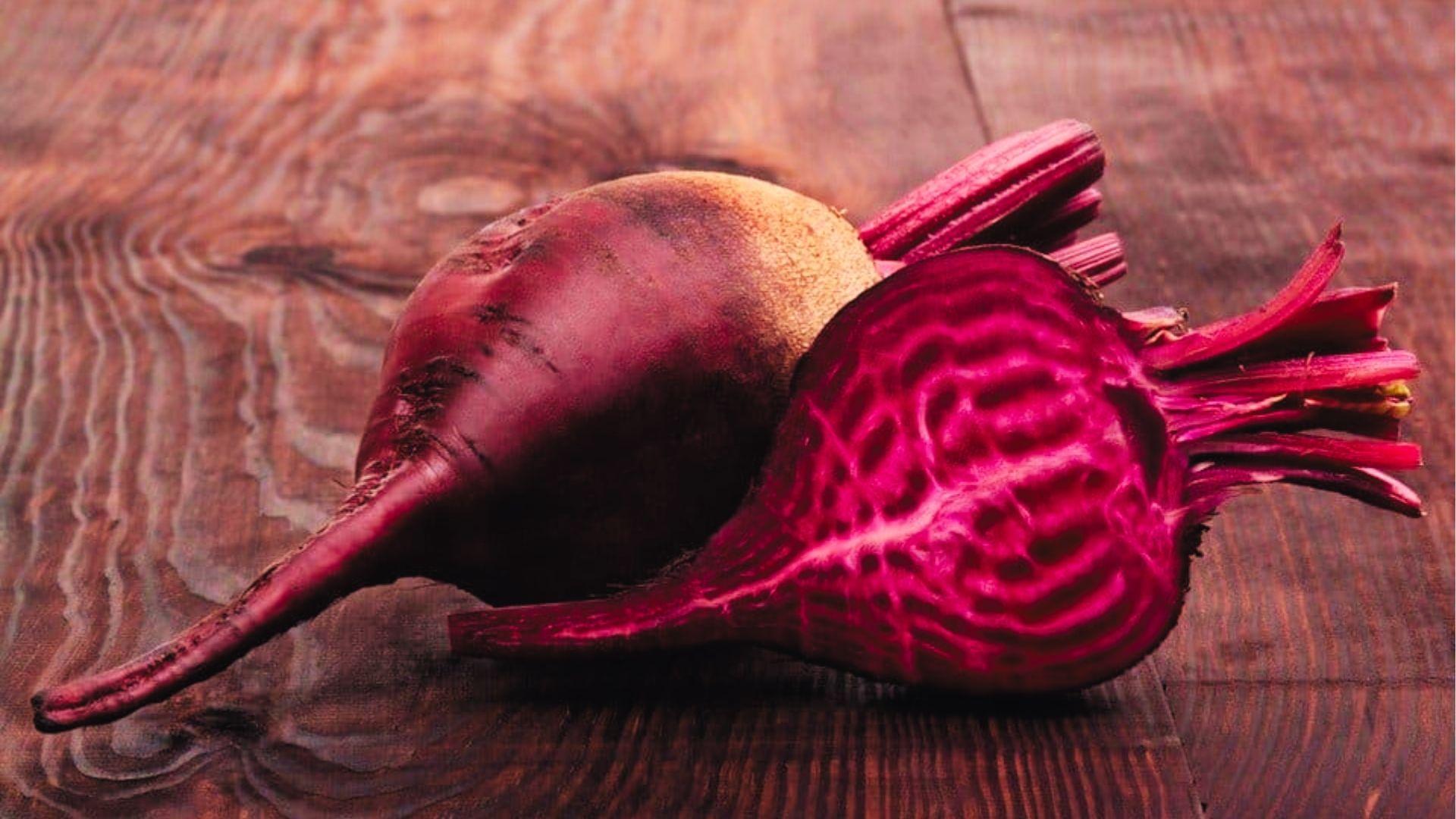 Whole and halved red beet on wooden surface revealing vivid magenta rings inside, earthy skin.